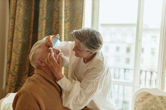 picture of elderly woman putting eyes drops on an elderly men sitting on a chair  