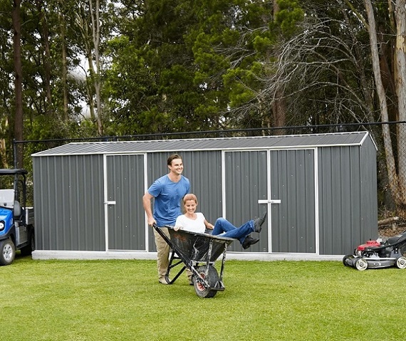 man pushing a woman in a wheelbarrow in front of a large storage shed 