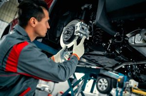 A mechanic in a grey and red uniform is installing a brake disc on a lifted vehicle in an auto repair shop.
