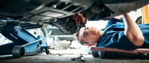 A mechanic in a blue uniform lying on a creeper under a car while working on its underside, with a hydraulic floor jack in the foreground.