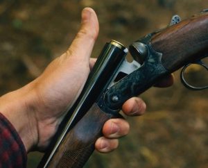 A close-up of a person inserting a shell into a break-action shotgun, with a forested background.