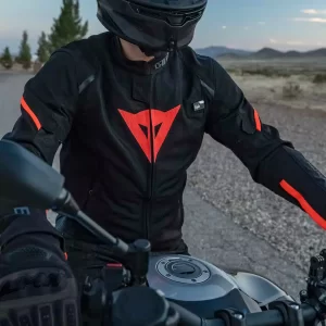 Motorcycle rider wearing a Dainese jacket with red accents, riding a bike on a road with mountains in the background.