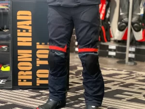 Close-up of a person wearing motorcycle pants with knee protection, standing in a showroom with a sign that says 'FROM HEAD TO TOE.