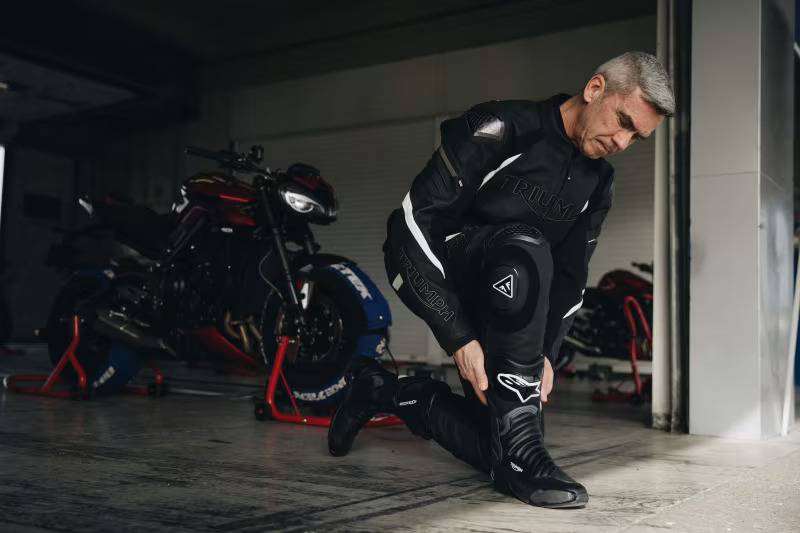 Motorcycle rider in a Triumph racing suit adjusting his gear in a garage with a Triumph motorcycle in the background.